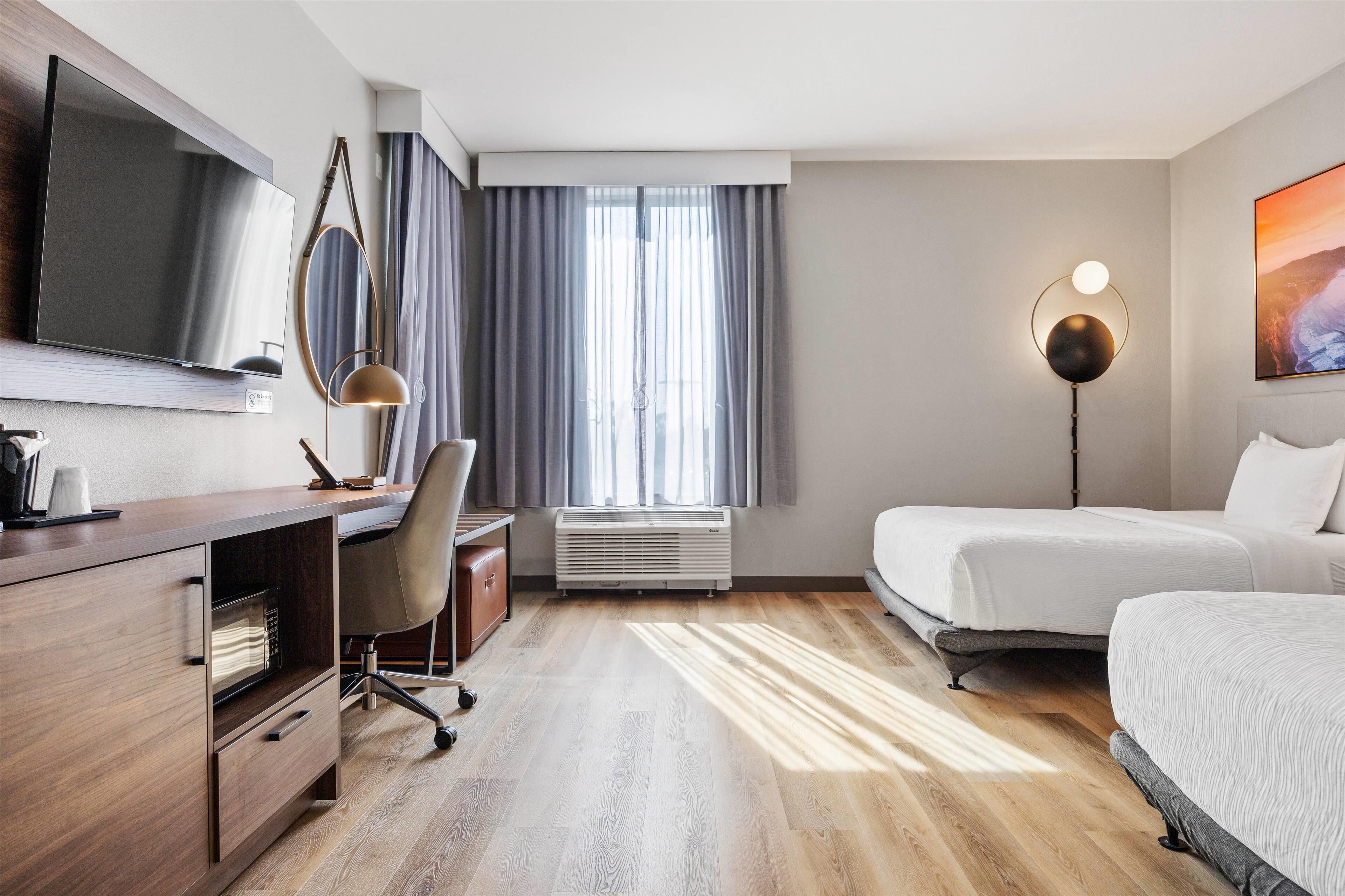 Habitación de hotel con televisor de pantalla plana, escritorio con silla, lámpara de pie, cama y una ventana grande con cortinas.