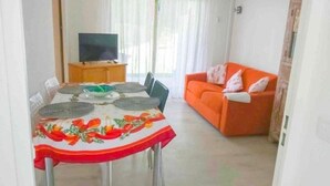 A dining area with a table set for four, a television on a stand, and an orange sofa in the background. - T2 A/C côte d'Azur, piscine, plage á 350m, WIFI  (Vallauris)