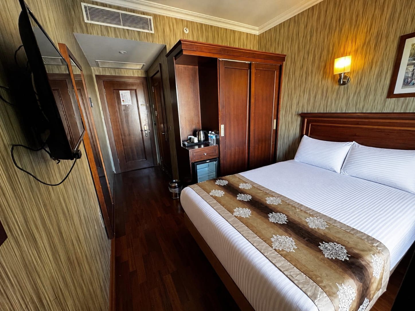 A hotel room with a bed, wooden wardrobe, a television, and a wall-mounted lamp.