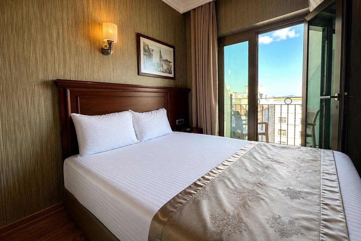 A hotel room with a large bed, a nightstand, a wall-mounted lamp, and a view of the outside balcony.