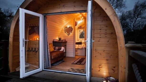 Una cabaña de madera acogedora y circular, con la puerta abierta que muestra un interior amoblado, incluyendo una cama y una lámpara con forma de corazón.