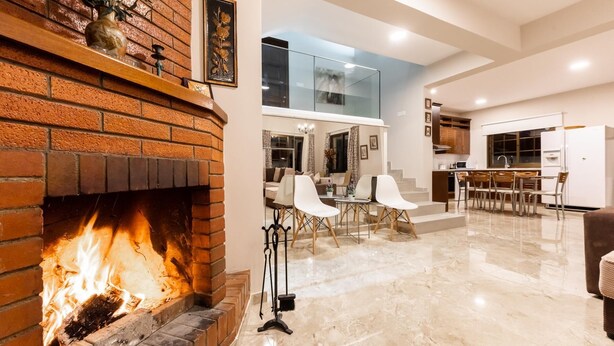 A modern living room with a brick fireplace, a dining area with white chairs, and a kitchen with white cabinets. - Chalet Pinecone by Ezoria Villas (Platres)
