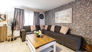 A living room with a sectional sofa, a wooden coffee table, a fan, a clock, and a framed picture on a brick wall.