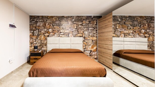 A bedroom with a stone wall, a wooden panel, a bed with a brown bedspread, a bedside table, and a nightstand.