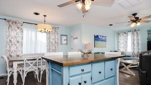 Un coin repas avec une table en bois et des chaises blanches, un meuble bleu avec des tiroirs et un ventilateur de plafond.