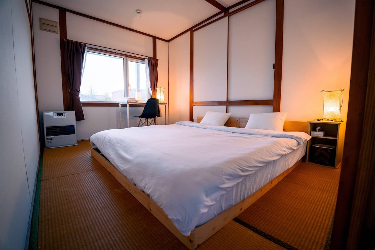 A hotel room with a large bed, a desk with a chair, a bedside table with a lamp, and a window with curtains.