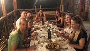 A group of people dining at an outdoor table with a white tablecloth, surrounded by stone walls and wooden beams. - Elizbar Talakvadze Winery (Gurjaani)