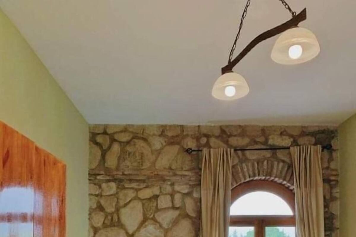 Une chambre avec un mur en pierre, un lit en bois, une fenêtre avec des rideaux, un radiateur et une table sur laquelle est posé un vase.