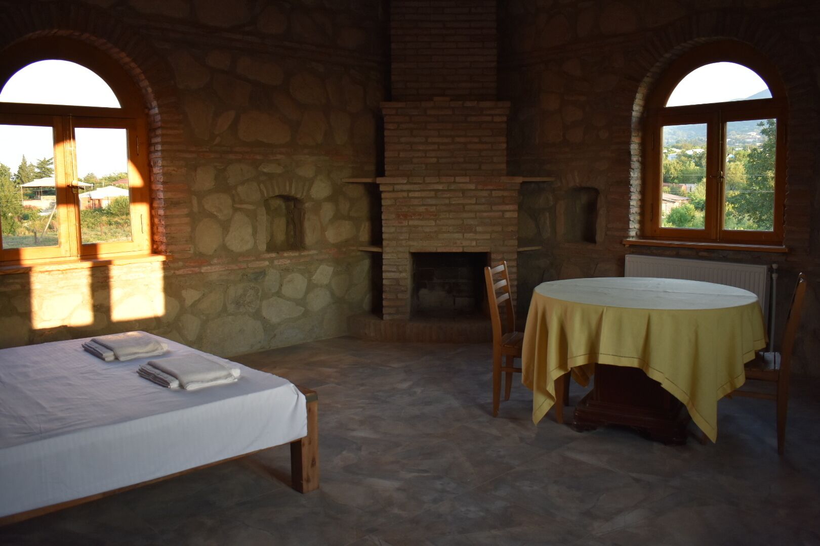 Una habitación rústica con chimenea de piedra, una cama con sábana blanca, una mesa redonda con mantel amarillo y dos ventanas de arco.