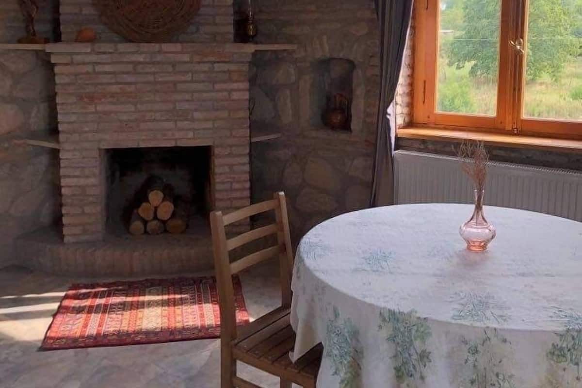 Une pièce avec une cheminée en pierre, une table ronde recouverte d’une nappe à motifs floraux et une fenêtre donnant sur un paysage verdoyant.