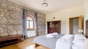 A bedroom with a stone wall, wooden floor, a bed with white linens, a wooden bench, and a wardrobe. - Elizbar Talakvadze Winery (Gurjaani)