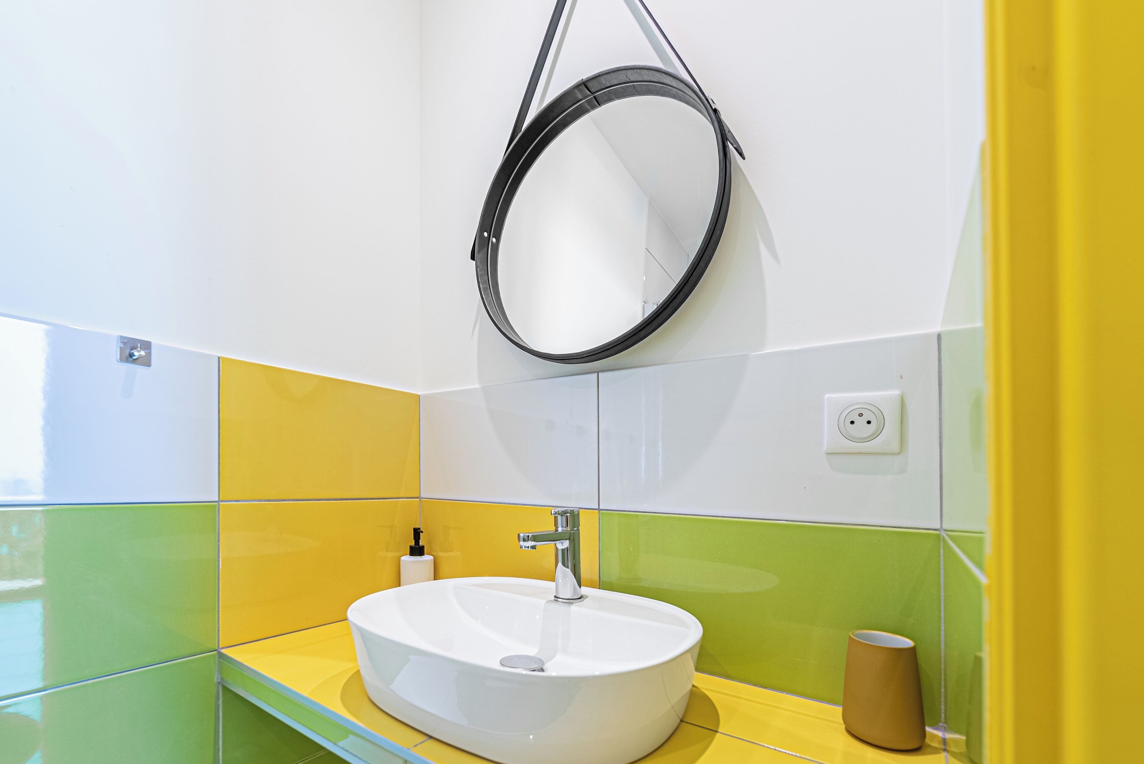 Een badkamer met een witte wasbak, gele en groene tegels, een ronde spiegel en een bruine beker op het aanrecht.