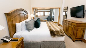 A hotel room with a wooden headboard, a bed with white linens, a fur throw, a wooden nightstand with a lamp and remote, a TV on the wall, and a view of the city through large windows. - Luxury Three Bedroom Suite with Three Kings 3 Suite (Park City)