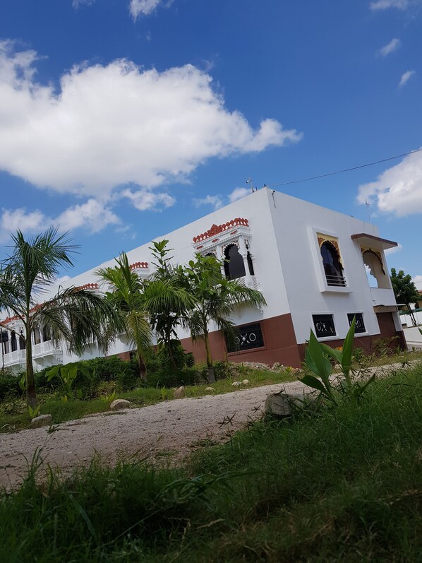 A white building with red accents and arched windows, surrounded by greenery and a clear sky. - Vijay Resort (Nathdwara)