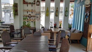 Un restaurant avec une longue table en bois et des chaises en osier, des étagères ornées d’objets décoratifs et des plantes en pot.