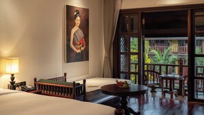 Une chambre d’hôtel avec un lit, une table de chevet avec une lampe, une chaise, une petite table avec des fruits, un tableau au mur et un balcon donnant sur un paysage verdoyant.