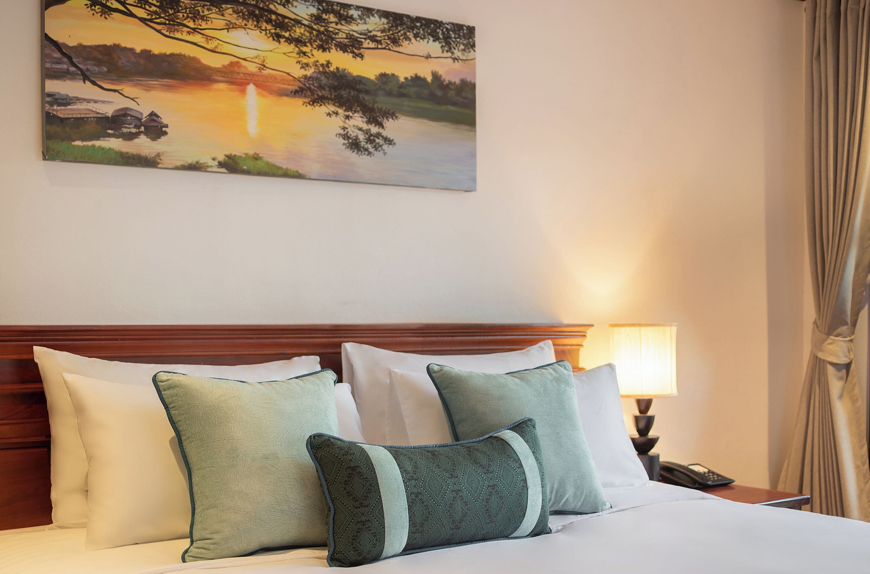 A hotel room with a bed, pillows, a nightstand, a lamp, and a painting of a sunset over a lake.