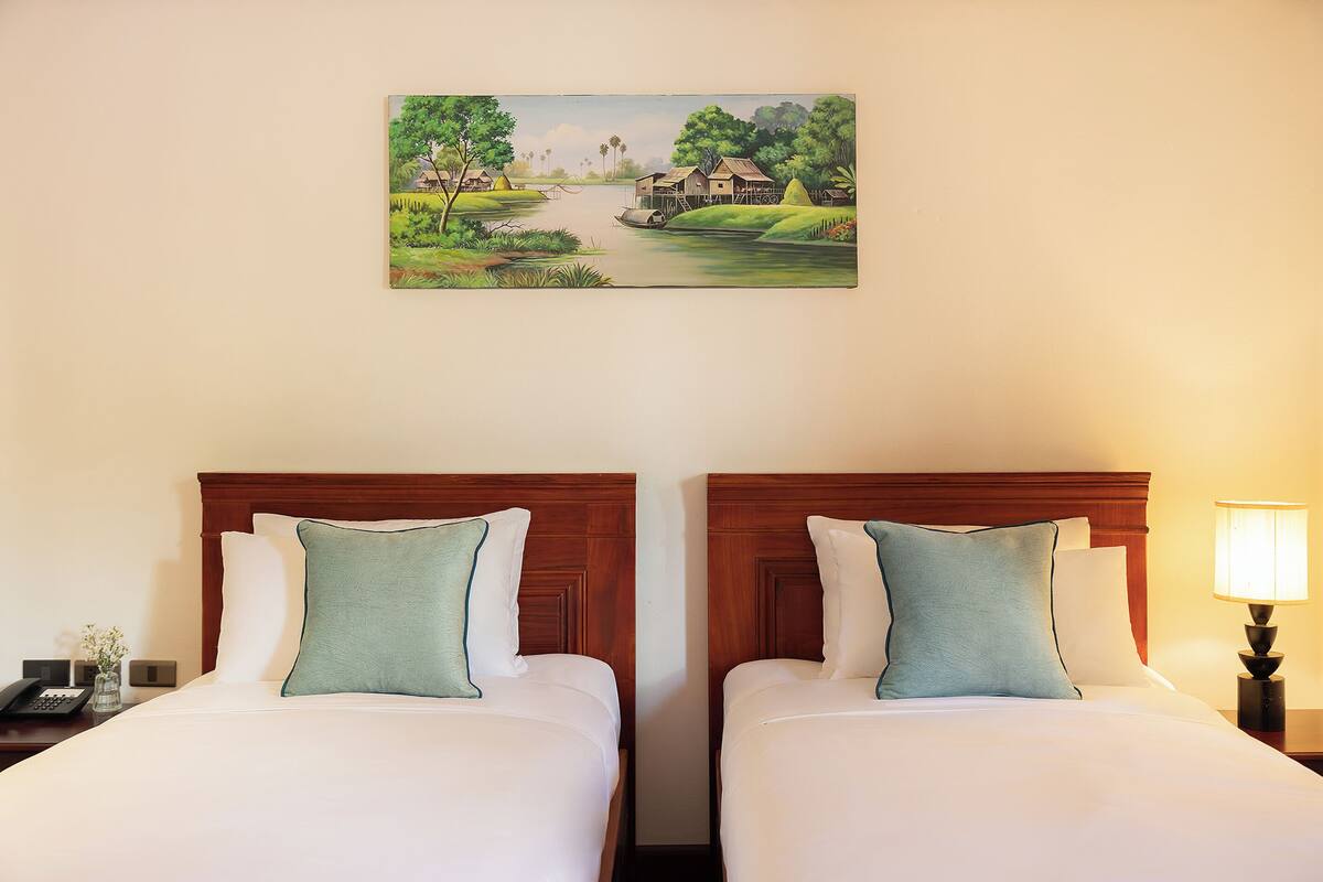 Habitación de hotel con dos camas, una mesita de noche con lámpara y un cuadro con una escena de río en la pared.