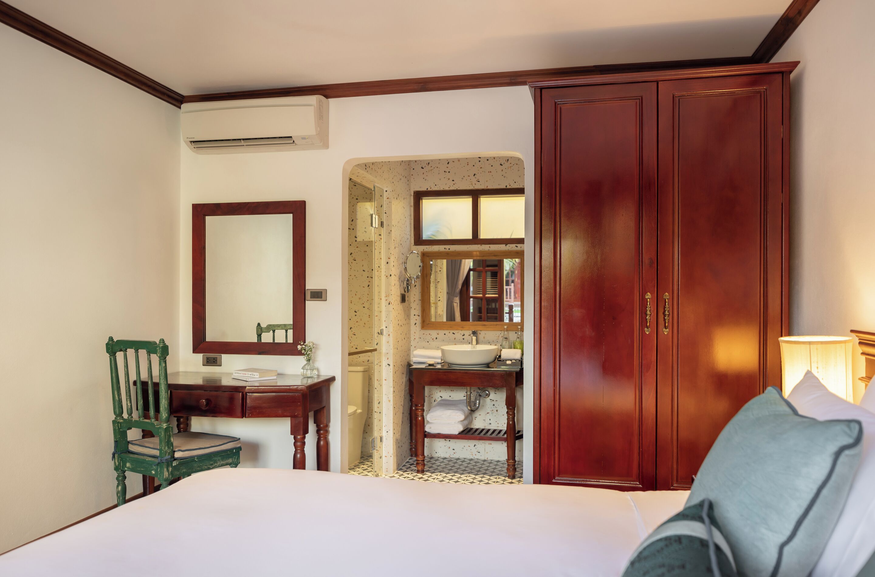 A hotel room with a bed, a wooden wardrobe, a desk with a chair, a bathroom with a sink and mirror, and a window.