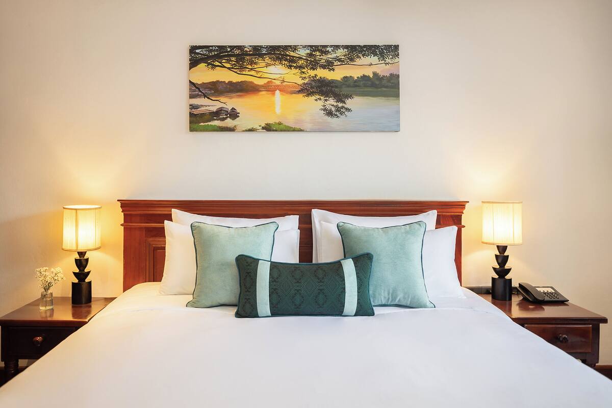 Una cama bien hecha con dos almohadas, una mesita de noche con una lámpara, y un cuadro de un atardecer sobre un lago en la pared.