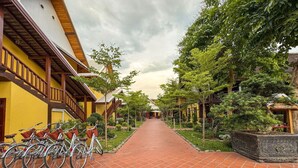 Un chemin bordé de vélos et de plantes en pot, qui mène à un bâtiment aux murs jaunes et bruns et au toit recouvert de tuiles.