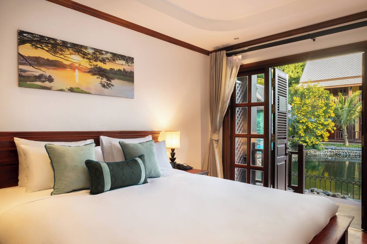 Habitación de hotel con una cama grande, un cuadro de un atardecer, vistas a un jardín y un balcón con un estanque.