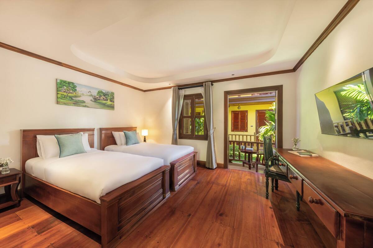 Habitación de hotel con dos camas de madera, un escritorio y un balcón con vistas a la vegetación.