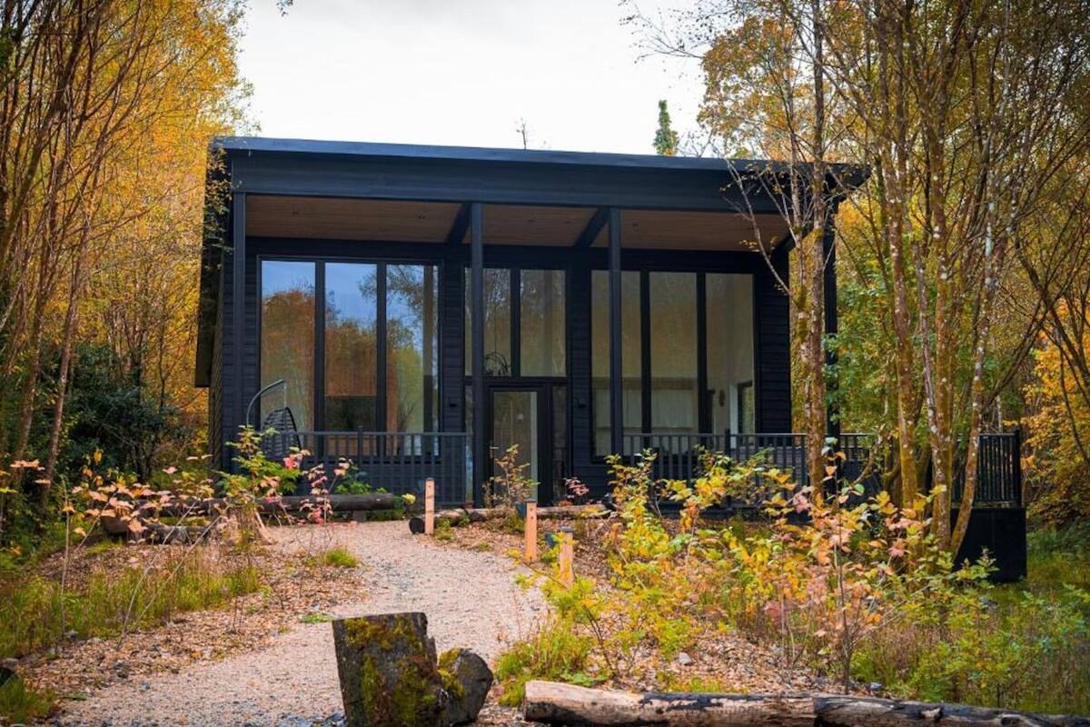 A modern building with large windows and a covered outdoor area, surrounded by trees and a gravel path.