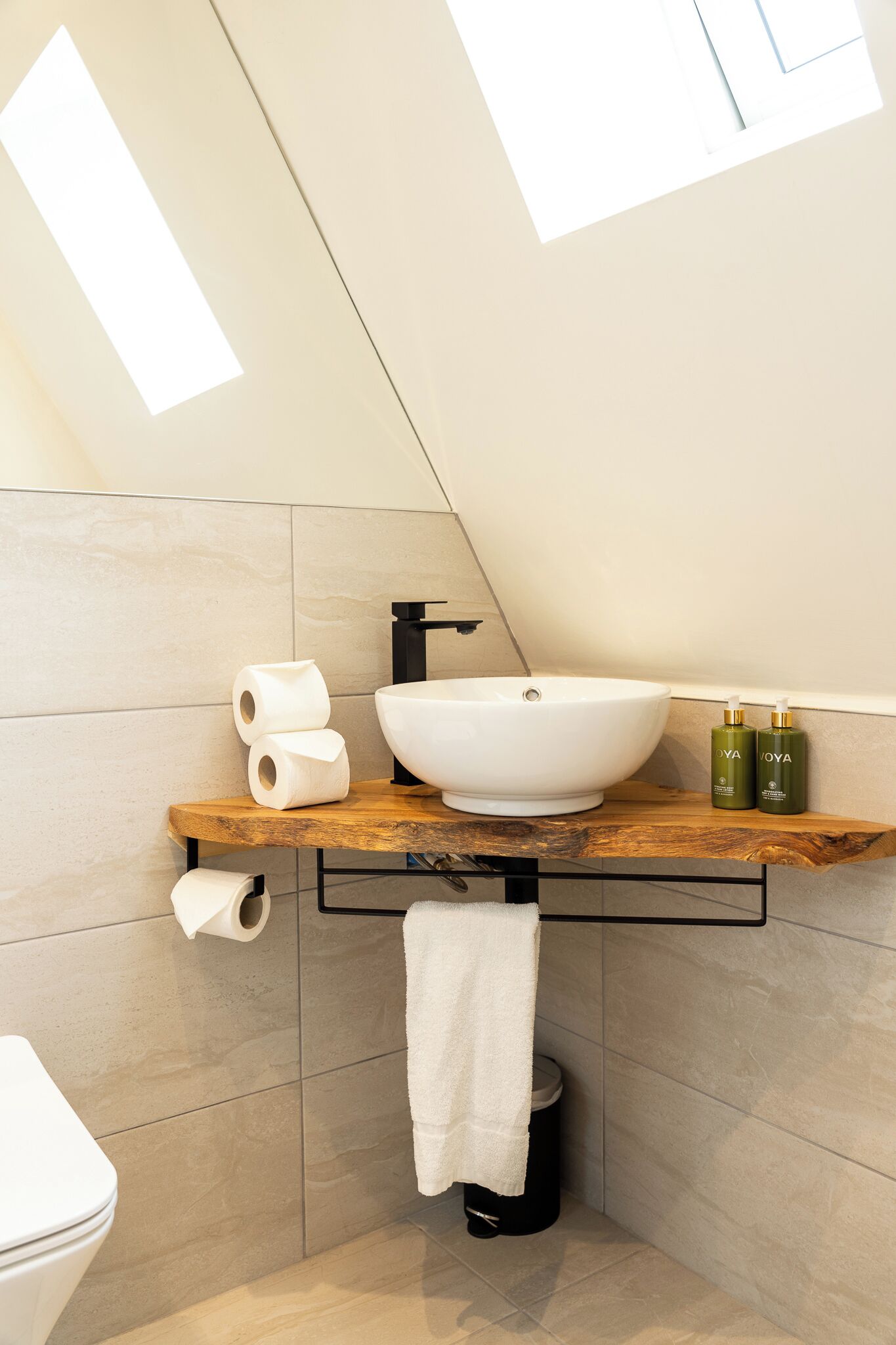 A modern bathroom with a white vessel sink, wooden countertop, toilet paper, and two green bottles.
