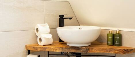 A modern bathroom with a white vessel sink, wooden countertop, toilet paper, and two green bottles.