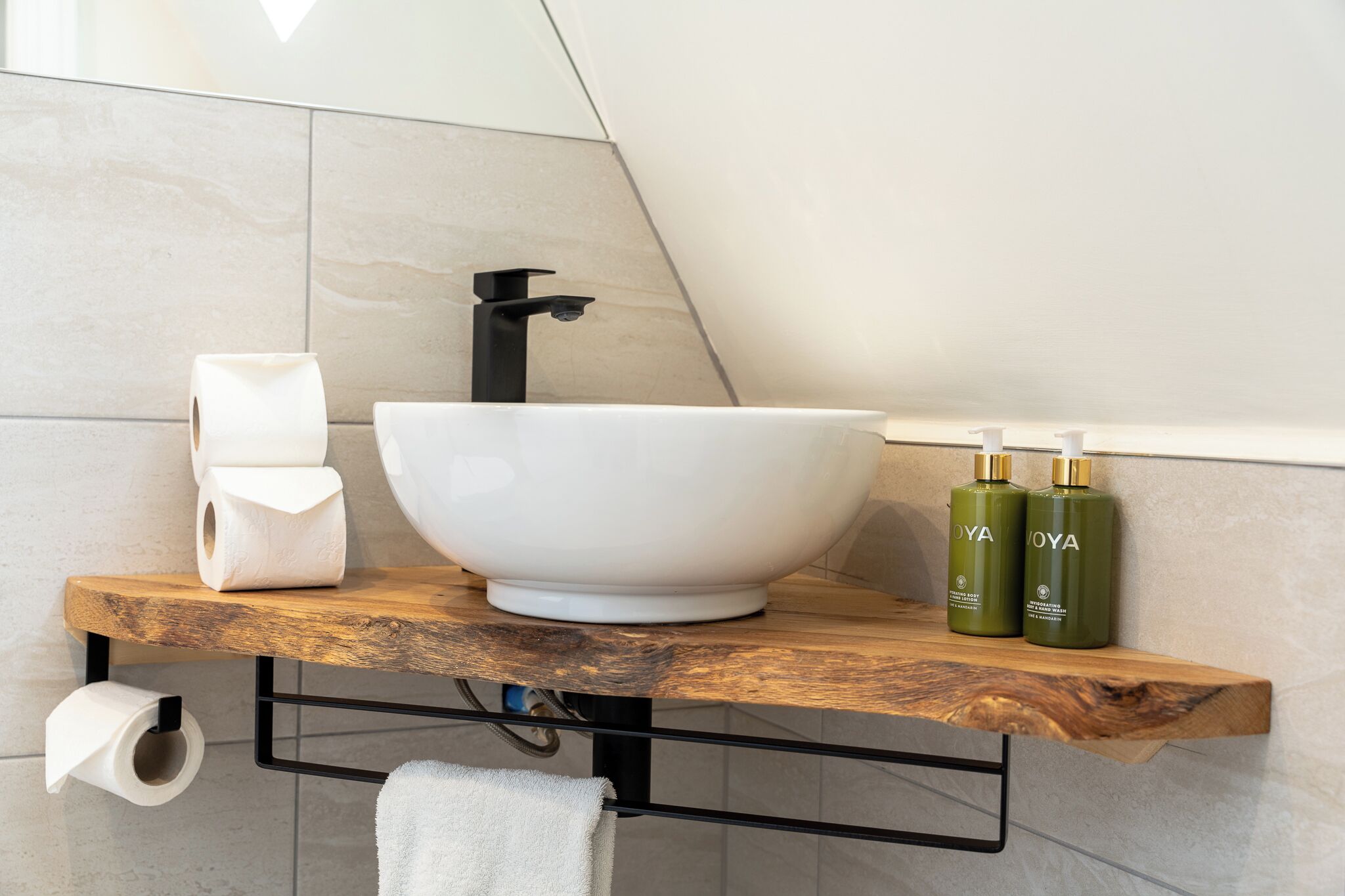 A bathroom with a large white vessel sink, a wooden shelf with toiletries, and a towel hanging on a rack.