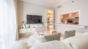 A modern living room with a flat-screen TV, a white sofa, a coffee table, and a kitchen area visible through an archway. - Chic Studio in Burj Views Podium, Downtown Dubai by Deluxe Holiday Homes (Dubai)