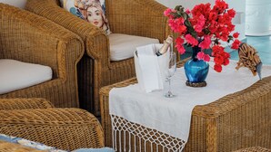 Un rincón de descanso de mimbre con una mesa blanca dispuesta para una comida, un jarrón de flores rojas y una botella de champán.