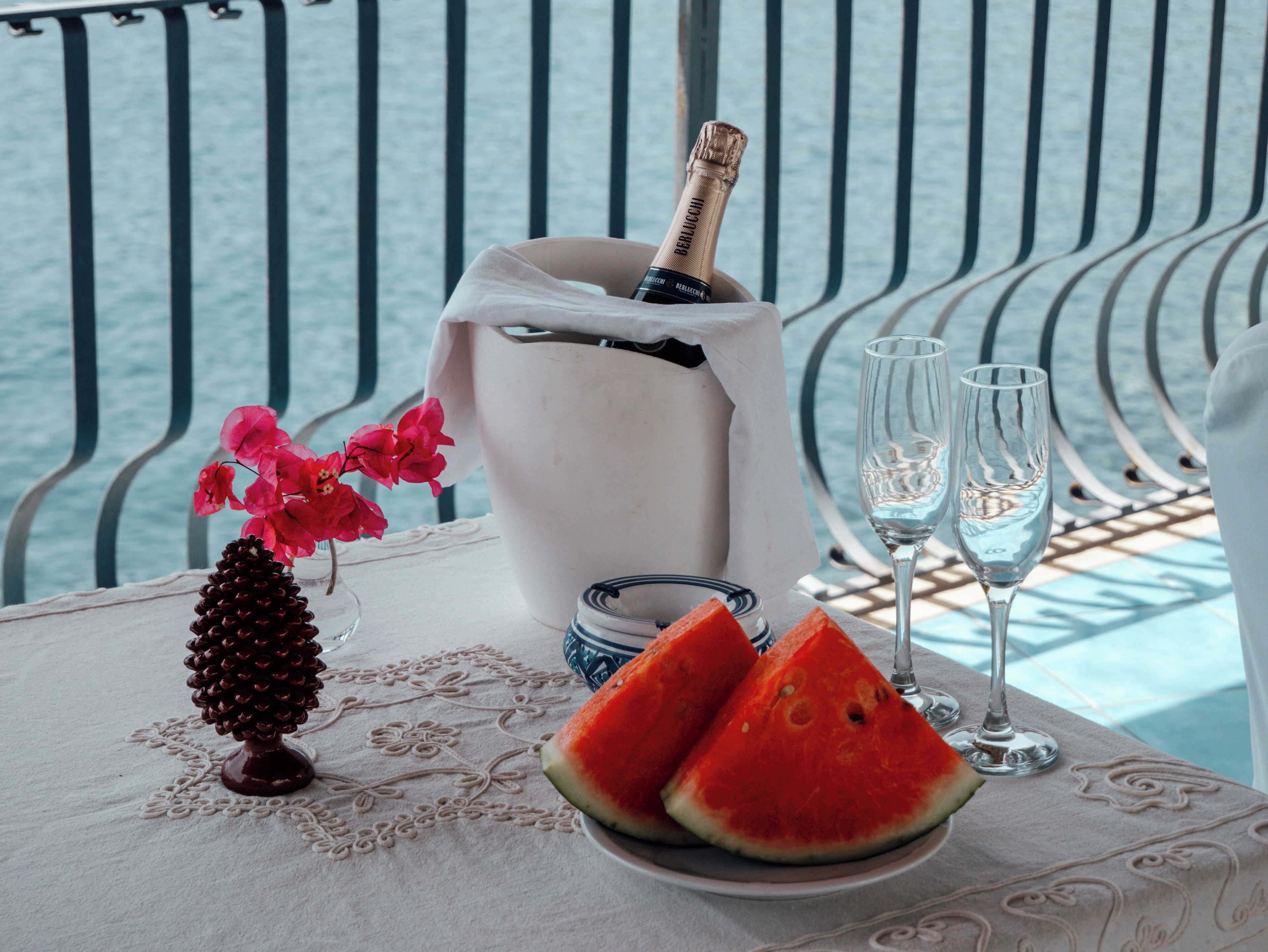 A table with a bottle of champagne, two glasses, a plate with watermelon slices, and a vase with flowers.