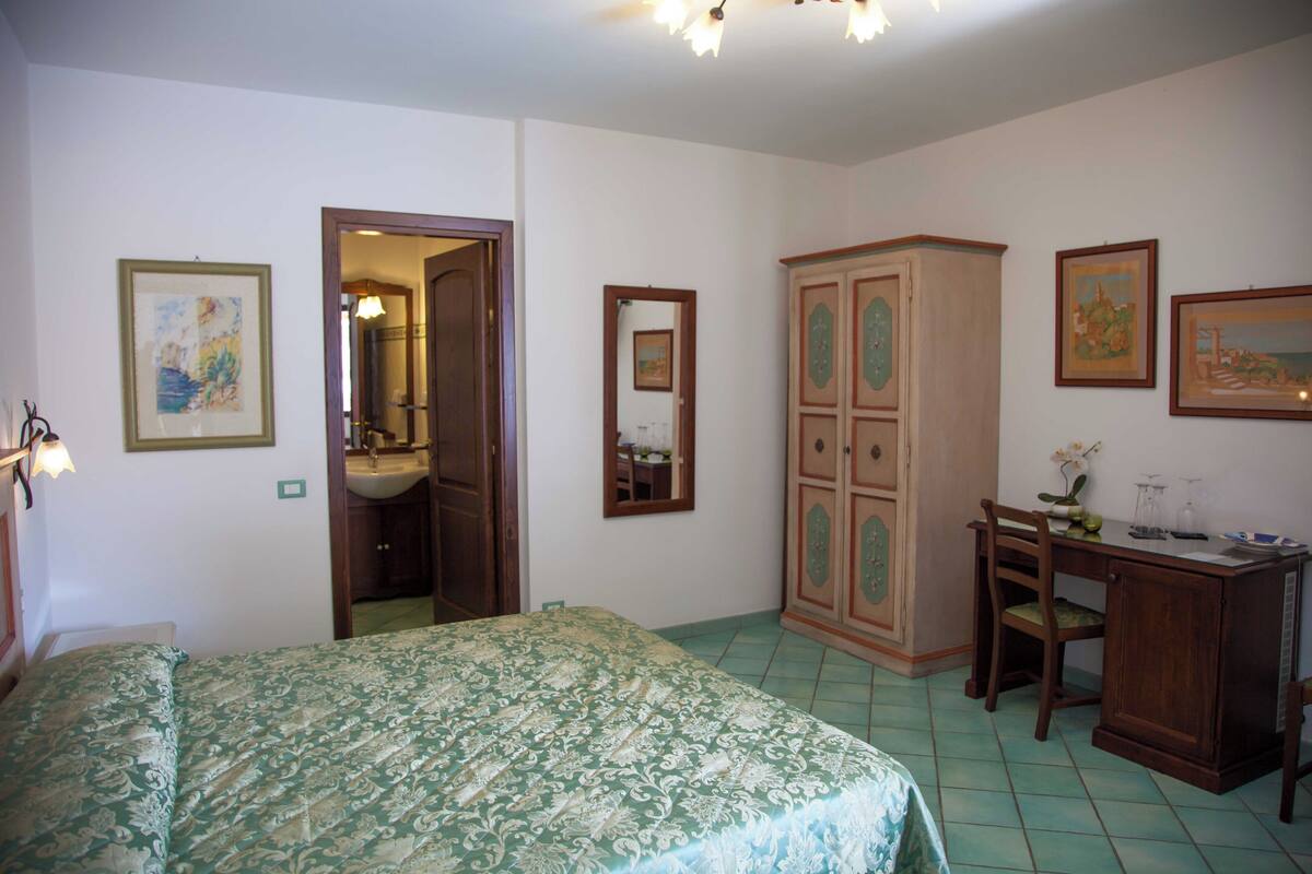 A bedroom with a bed, a desk, a wardrobe, and a bathroom visible through an open door.