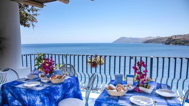 Una mesa de comedor dispuesta con platos, pan y flores en un balcón con vista al mar.