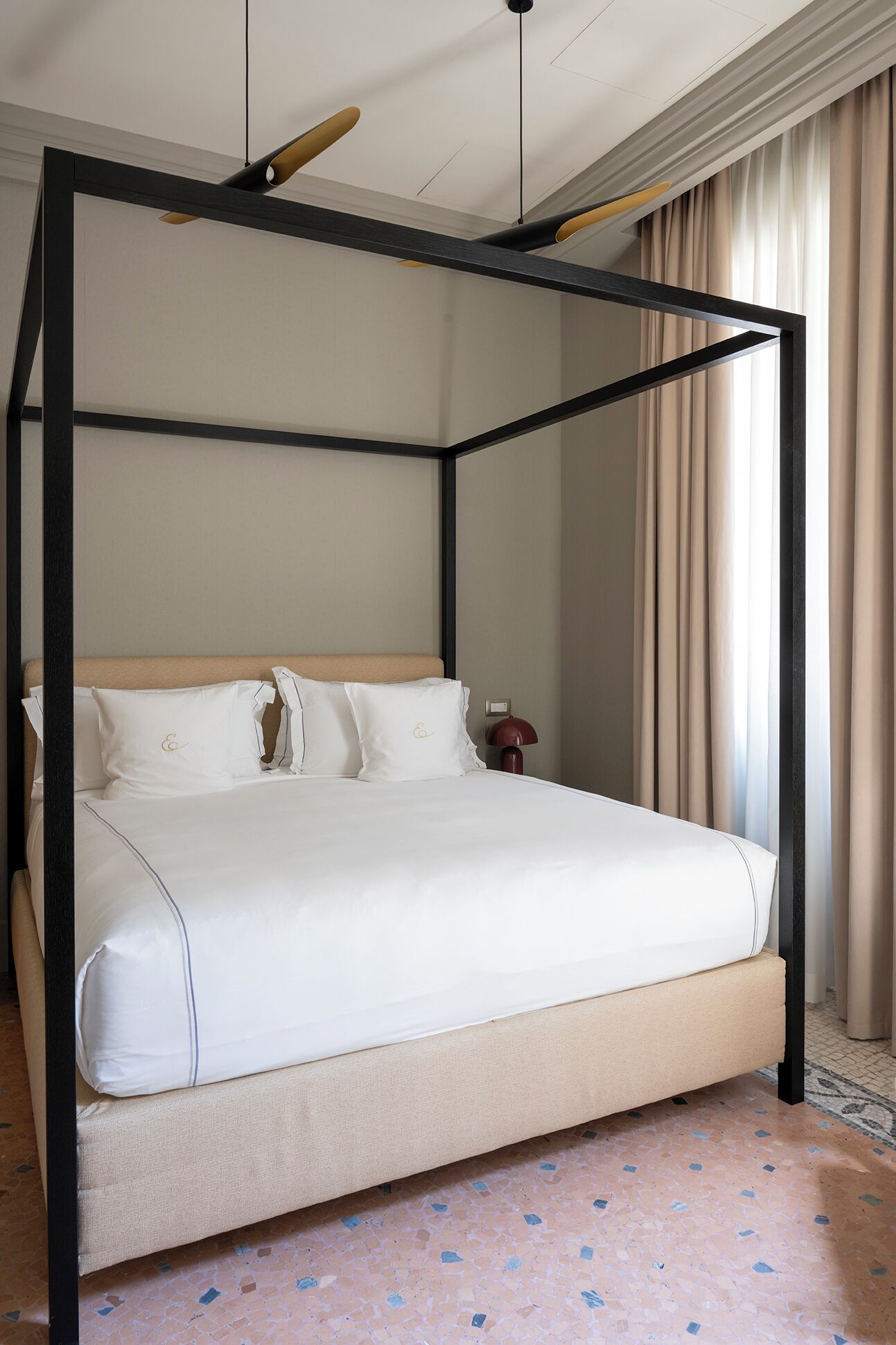 Una cama con dosel, un cabecero color beige y ropa de cama blanca, una mesita de noche con una lámpara, y vistas a una ventana con cortinas.