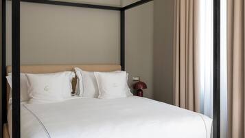 Una cama con dosel, un cabecero color beige y ropa de cama blanca, una mesita de noche con una lámpara, y vistas a una ventana con cortinas.