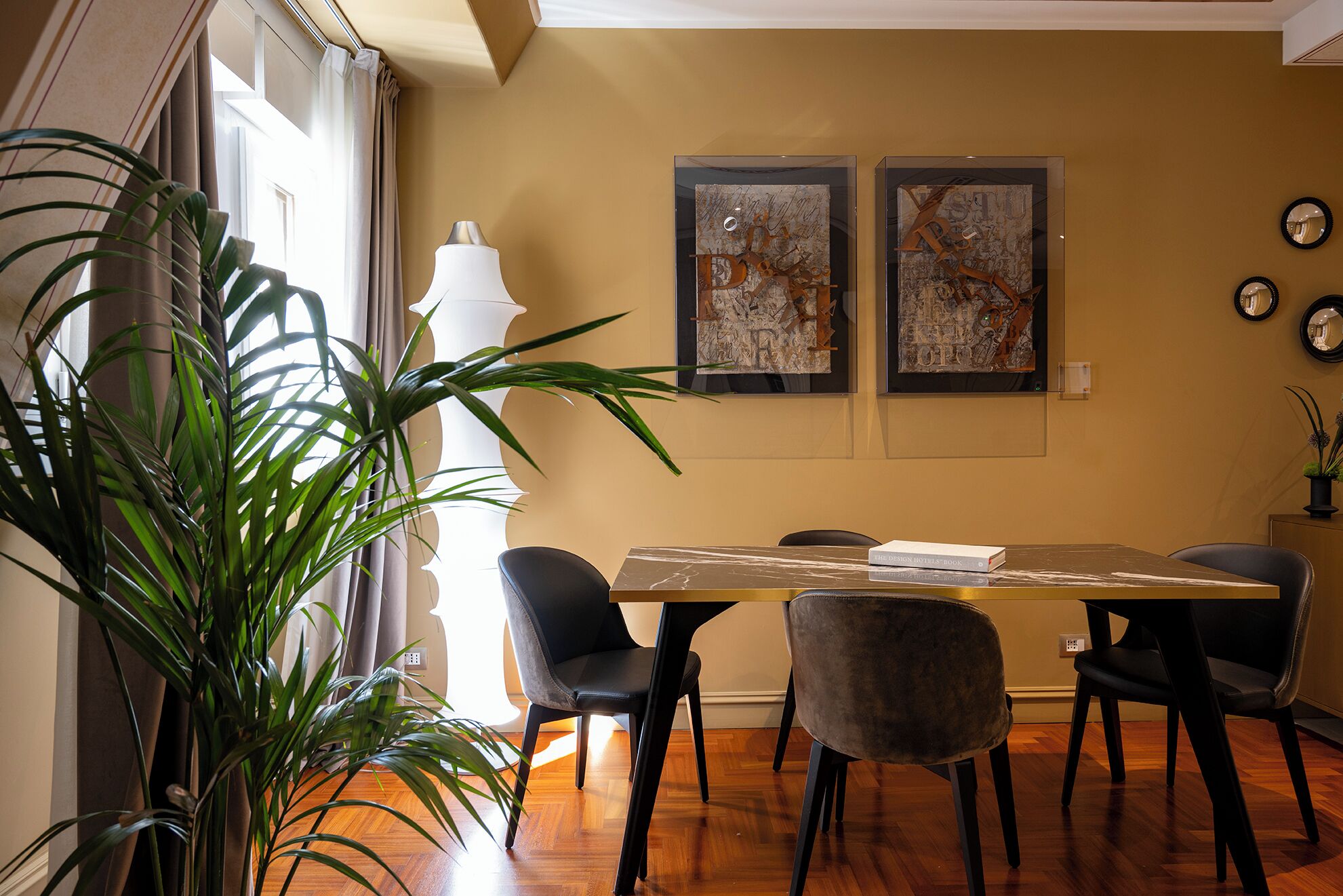 Un comedor con una mesa de madera, cuatro sillas, dos cuadros abstractos, una planta en maceta y una lámpara.
