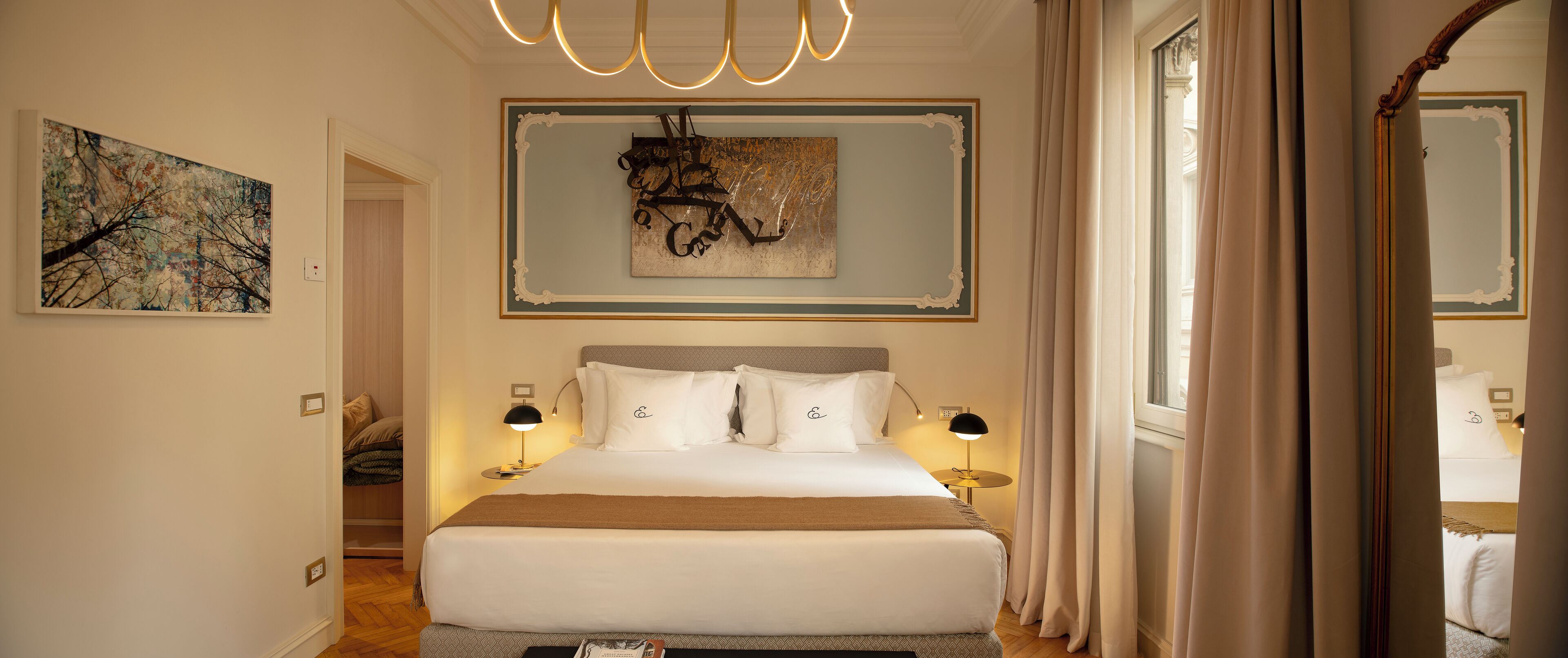 A well-lit hotel room with a large bed, two bedside tables with lamps, a painting on the wall, and a view through a window with curtains.