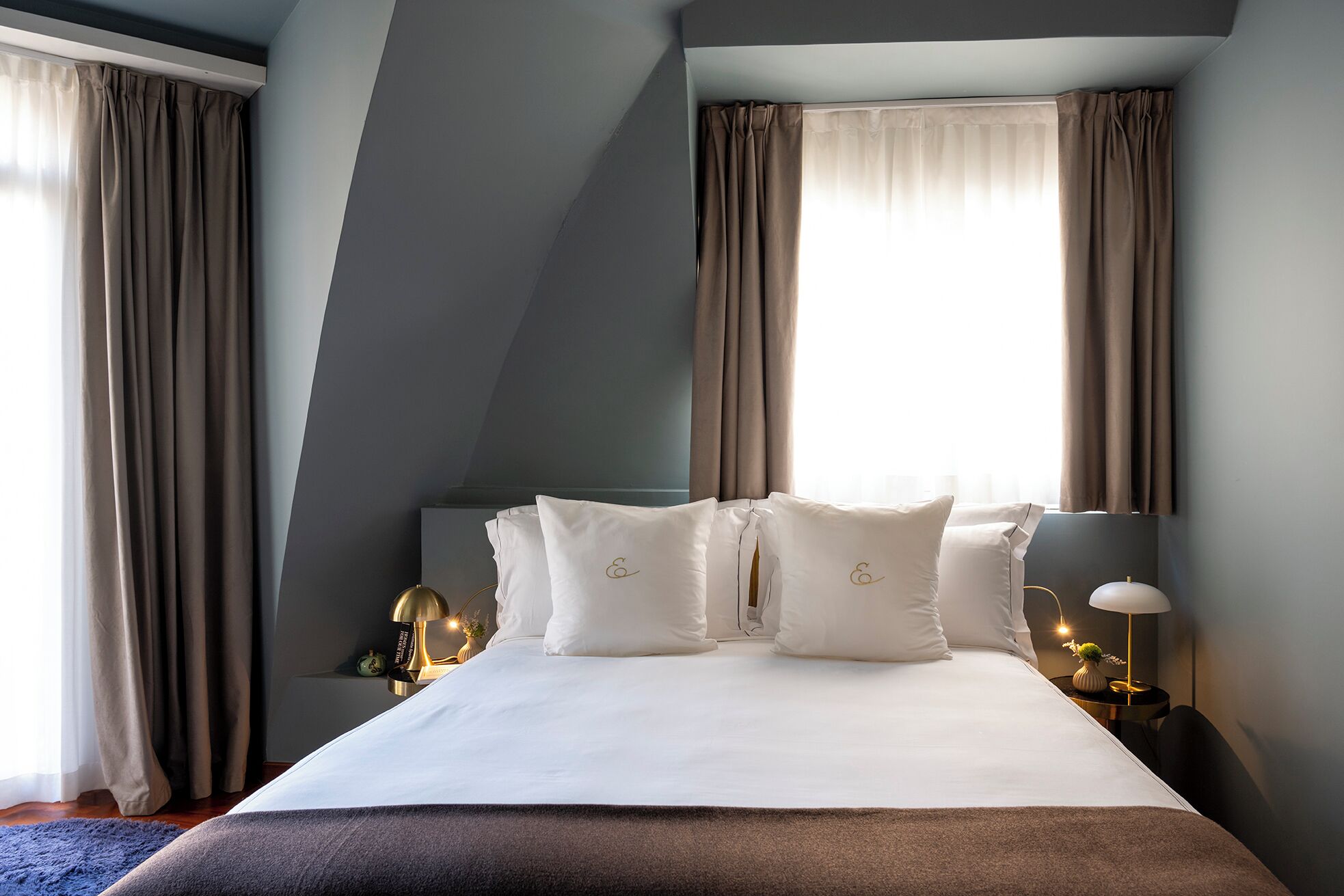 Una habitación de hotel moderna con una cama grande, dos mesitas de noche y un ventanal con cortinas.