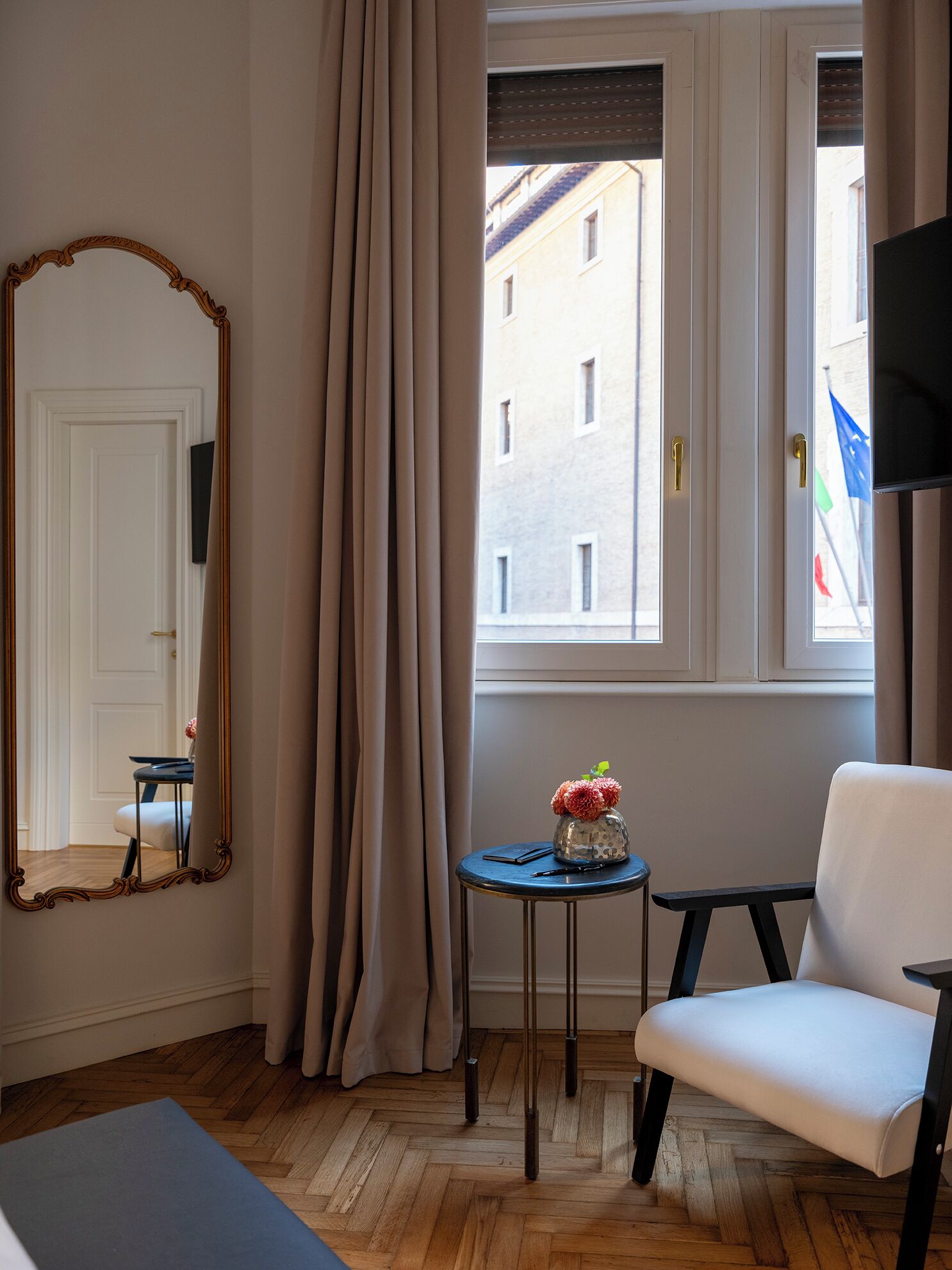 A room with a large mirror, a small table with a vase, a chair, and a window with a view of buildings.