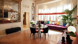 Un'ampia stanza con un grande specchio decorato, un tavolo rotondo con sedie, un divano con cuscini colorati e un'ampia finestra con tende leggere.