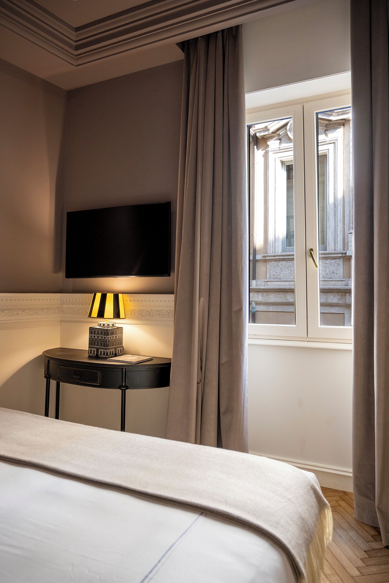 A hotel room with a bed, a bedside table with a lamp, a flat-screen TV mounted on the wall, and a window with curtains.