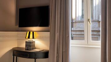 A hotel room with a bed, a bedside table with a lamp, a flat-screen TV mounted on the wall, and a window with curtains.