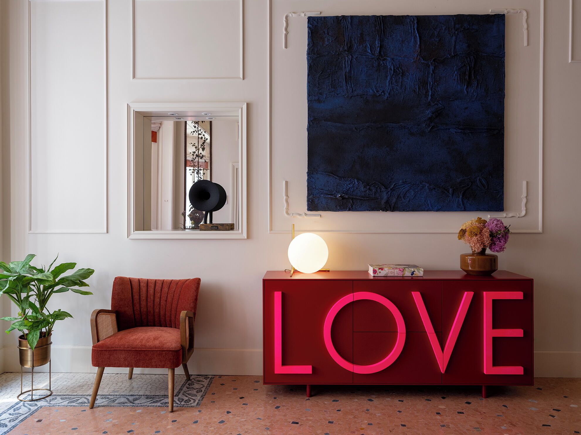 Una habitación con un letrero rojo que dice LOVE, un cuadro azul oscuro grande, una silla roja, una planta en maceta y un piso con diseño de mosaico.