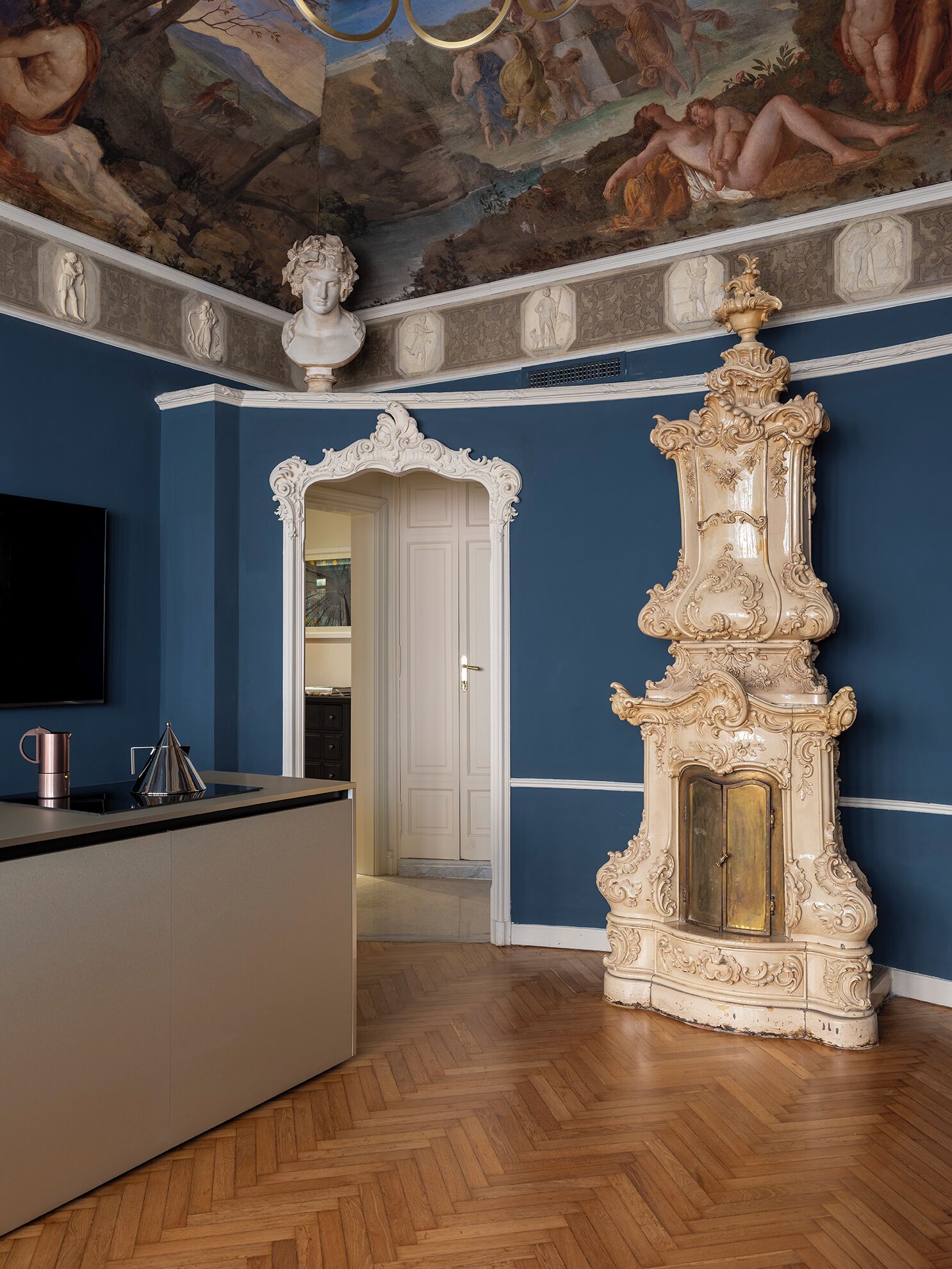 A room with a blue wall, a wooden herringbone floor, a white ornate fireplace, and a flat-screen TV mounted on the wall.