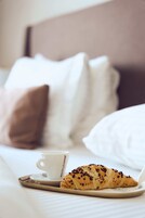 Una bandeja con un croissant y una taza de café sobre una cama con ropa blanca y una almohada marrón.