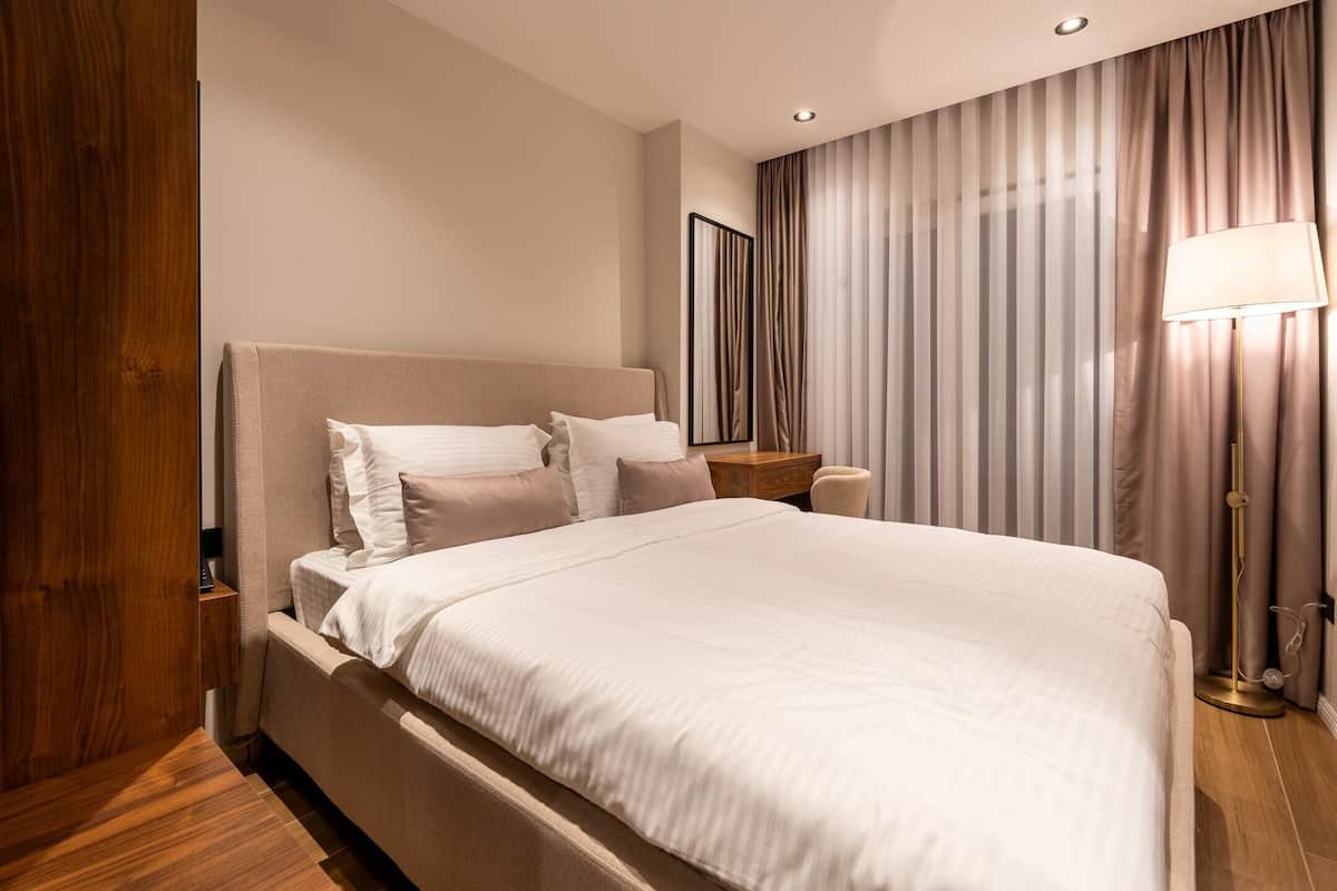 A modern hotel room with a large bed, a bedside table, a lamp, and a view of the outside through curtains.