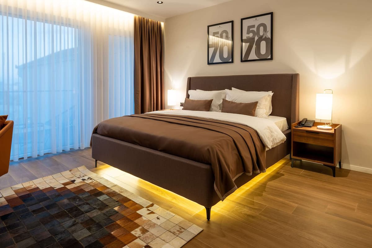A modern hotel room with a large bed, a nightstand, a lamp, and a view of the outdoors through sheer curtains.